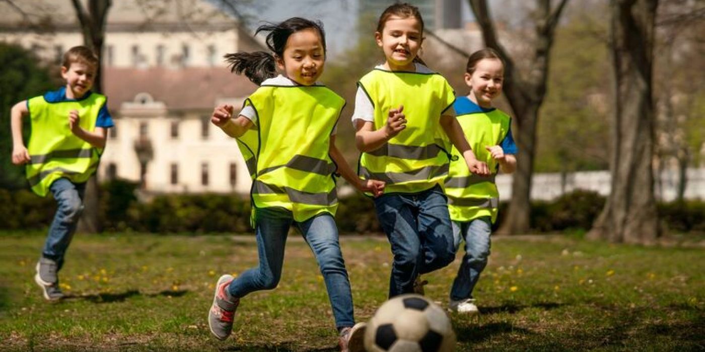 Fire barn løper etter en fotball på en gresslette i en park. Barna har på seg gule refleksvester over sine vanlige klær. De ser ut til å være i alderen 7-10 år og inkluderer både gutter og jenter med ulik etnisk bakgrunn.