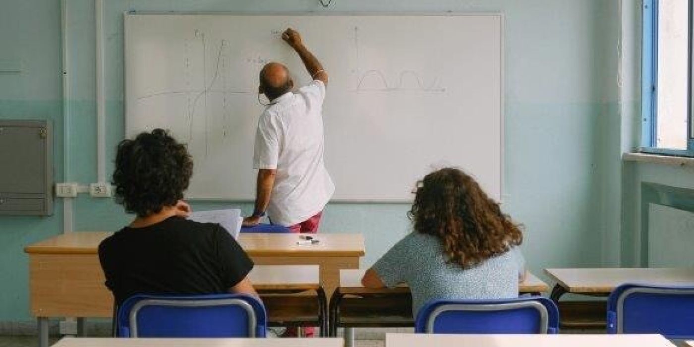 Lærer som skriver på tavle mens to voksne elever følger. Er