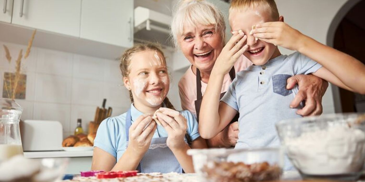 En eldre kvinne med hvitt hår står i et kjøkken sammen med to barn, en jente og en gutt. Alle tre ler og smiler bredt. På kjøkkenbenken foran dem er det ingredienser og kjøkkenutstyr, som antyder at de lager noe sammen.