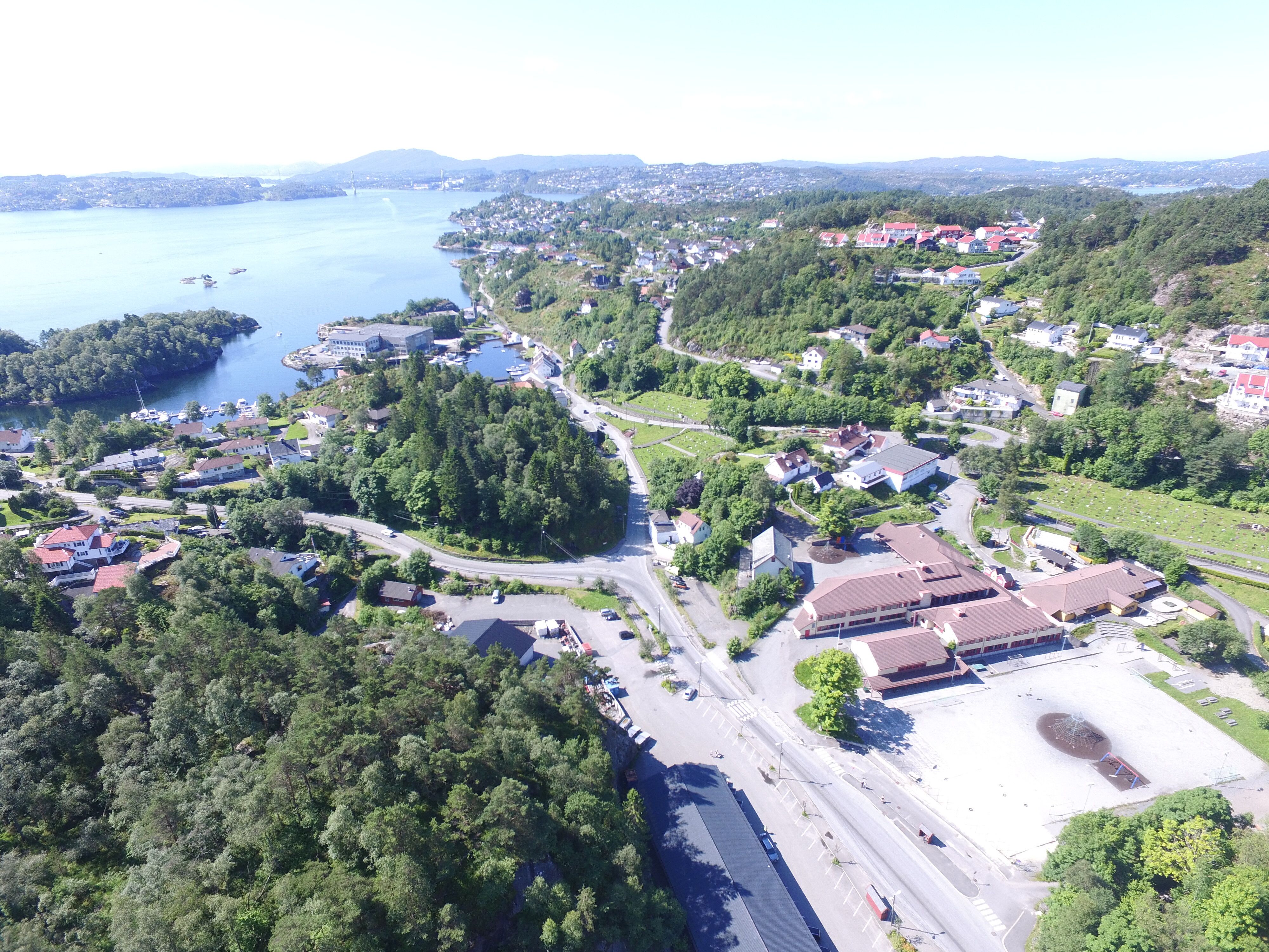 Luftfoto av en kystby i et fjordlandskap. Bildet viser en blanding av bebyggelse og frodig natur. I forgrunnen ses veier, parkeringsplasser og noen større bygninger med røde tak. Bebyggelsen strekker seg oppover grønne åser med skog. En stor fjord dominerer bakgrunnen, med flere øyer og landtunger synlige. I det fjerne skimtes fjell. Byen ser ut til å være godt integrert i det naturskjønne landskapet, med hus og veier som snor seg mellom trærne og langs vannkanten.