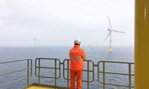 Mann i oransje arbeidsklær og hjelm med ryggen til, står og ser utover havet der det er flere vindmøller