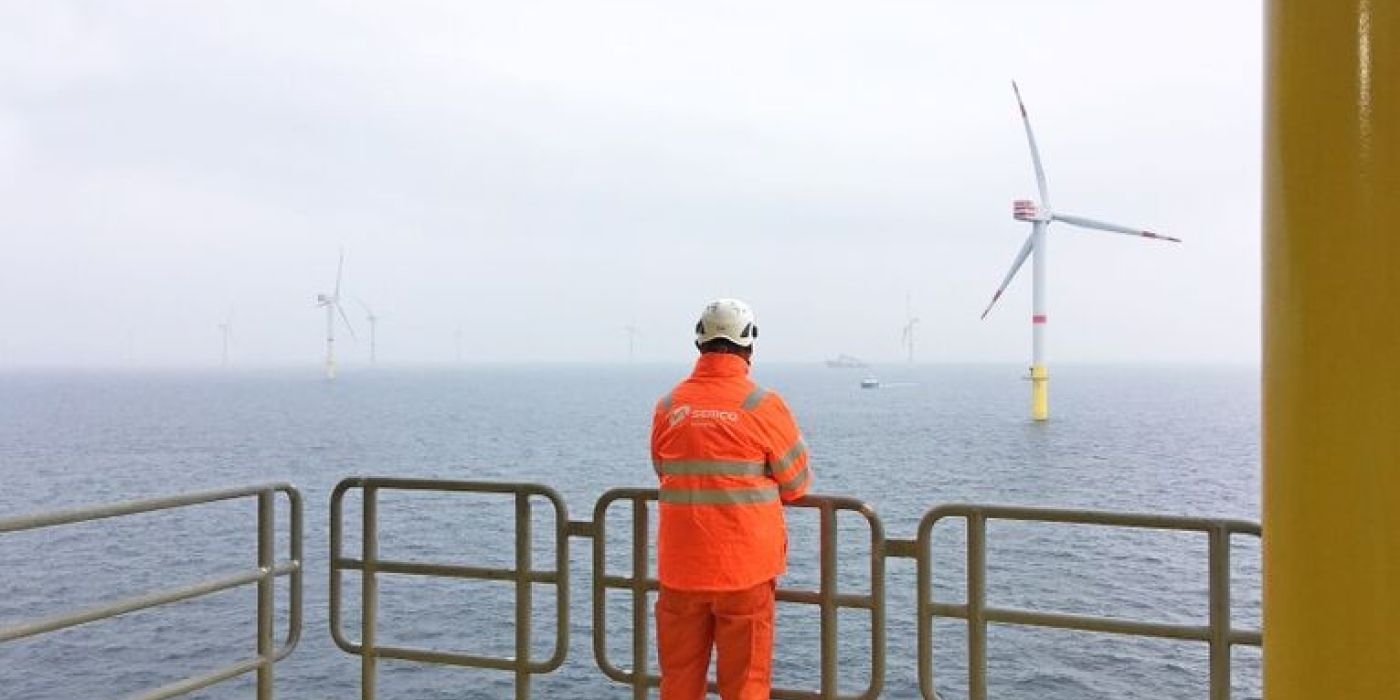 Mann i oransje arbeidsklær og hjelm med ryggen til, står og ser utover havet der det er flere vindmøller