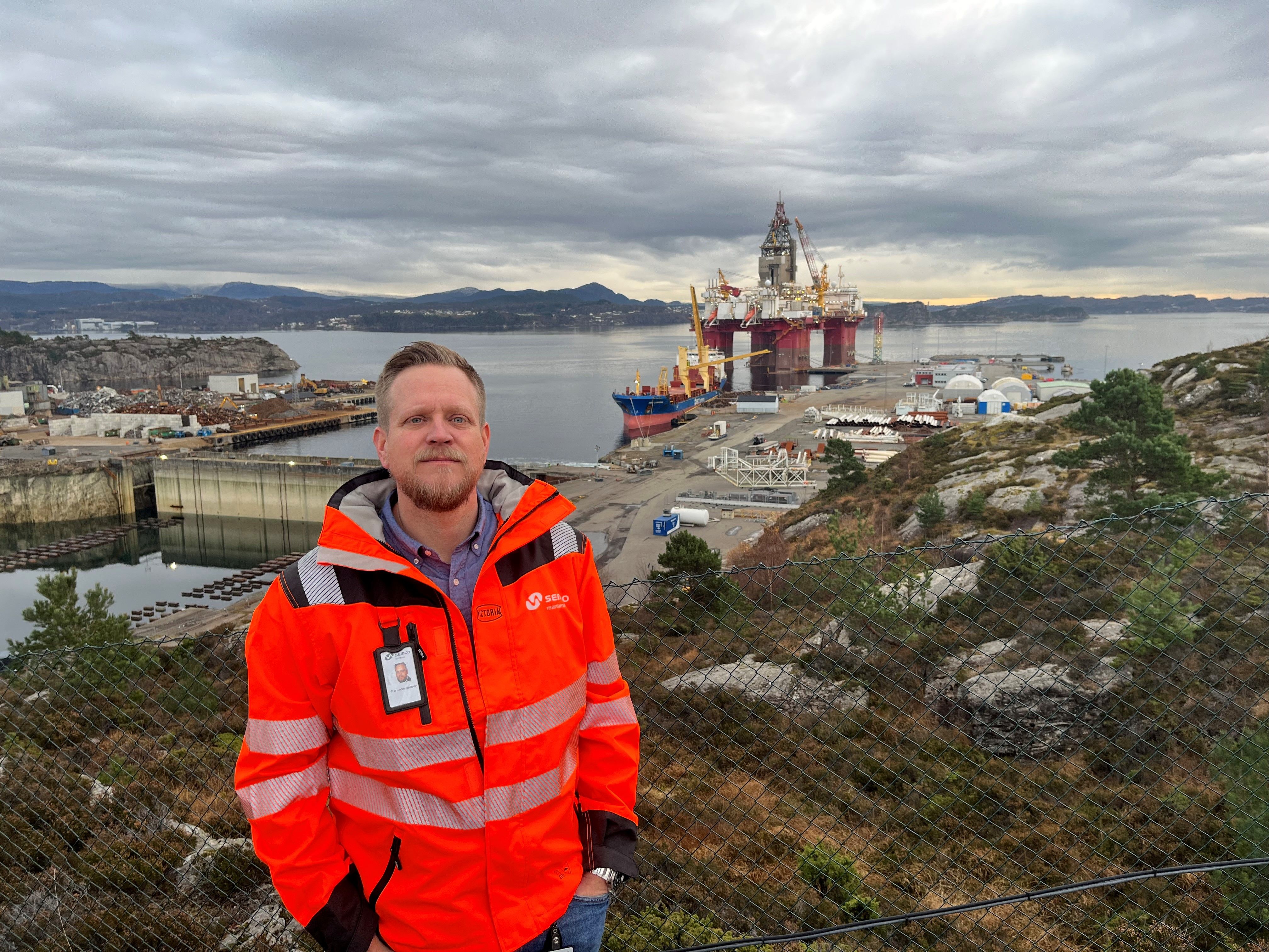 Mann i oransj jakke som står foran et industriområde og ser i kamera.