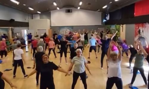 En stor gruppetrening i en romslig innendørs treningssal. Rommet er fylt med mennesker, hovedsakelig eldre voksne, som deltar i en aerobic-økt.