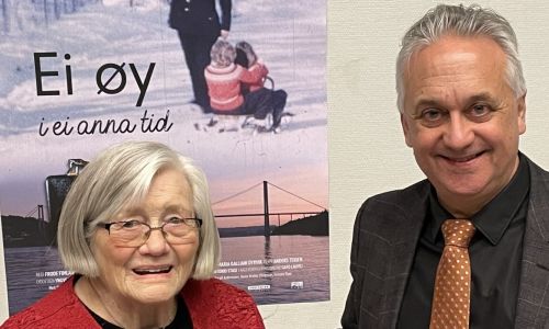 Bildet viser to personer stående foran en plakat med tittelen "Ei øy i ei anna tid". Personen til venstre er en eldre kvinne med grått hår og briller, iført en rød topp. Personen til høyre er en mann med grått hår, iført dressjakke, sort skjorte og oransje slips med mønster. Begge smiler mot kameraet. Bakgrunnen domineres av filmplakaten, som viser en vinterlig scene med en person som trekker et barn på akebrett.