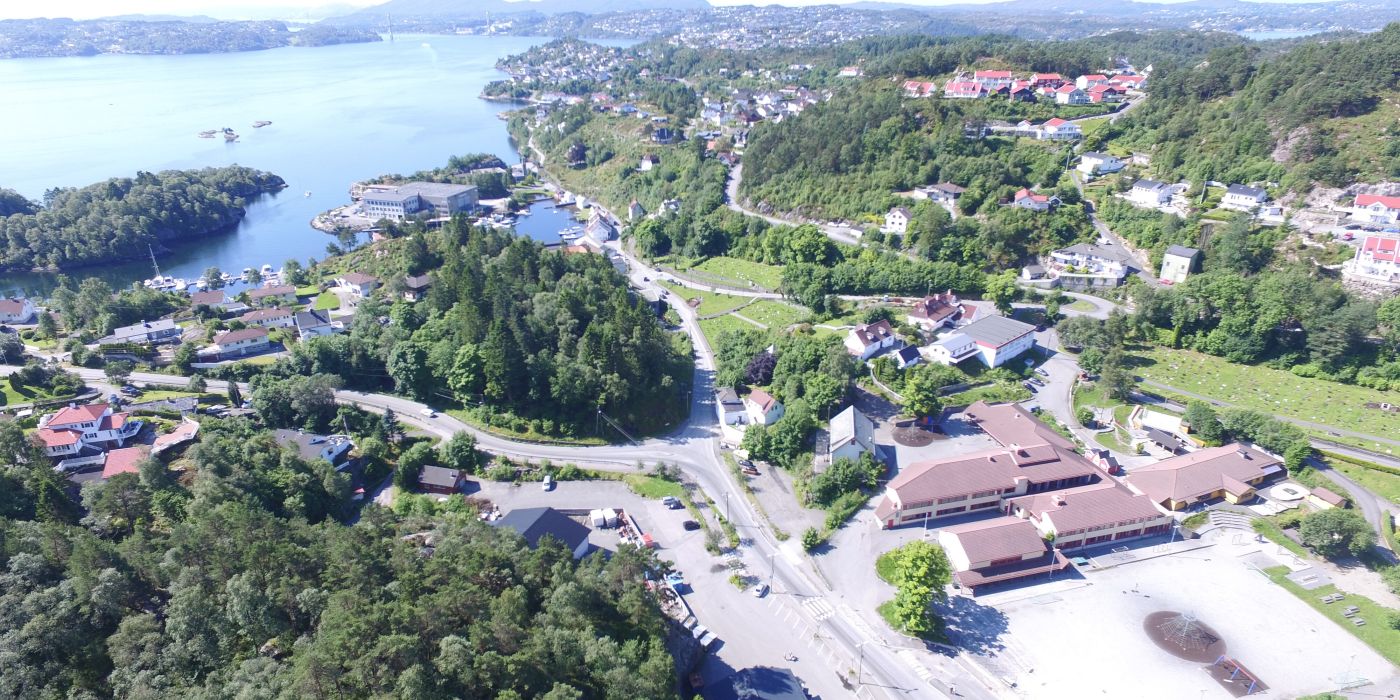 Luftfoto av en kystby i et fjordlandskap. Bildet viser en blanding av bebyggelse og frodig natur. I forgrunnen ses veier, parkeringsplasser og noen større bygninger med røde tak. Bebyggelsen strekker seg oppover grønne åser med skog. En stor fjord dominerer bakgrunnen, med flere øyer og landtunger synlige. I det fjerne skimtes fjell. Byen ser ut til å være godt integrert i det naturskjønne landskapet, med hus og veier som snor seg mellom trærne og langs vannkanten.