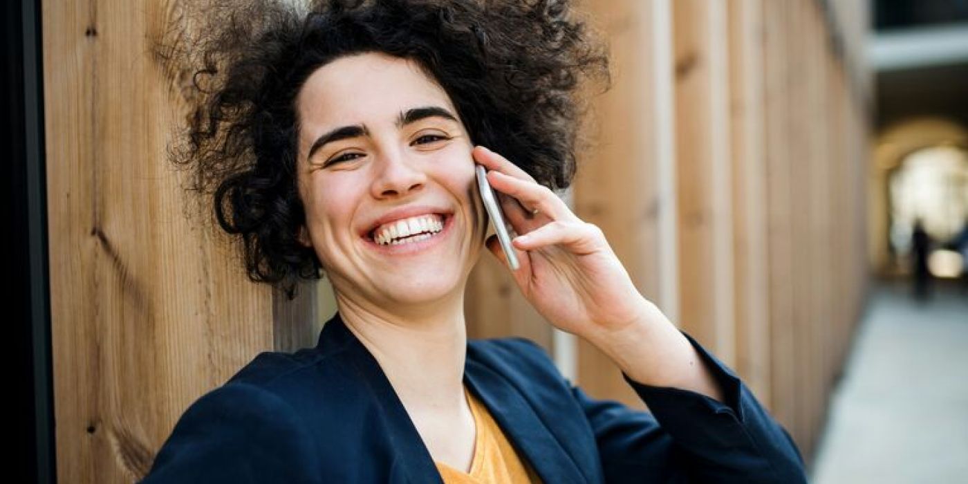 En ung kvinne med krøllete mørkt hår smiler bredt mens hun snakker i mobiltelefon. Kvinnen ser glad og avslappet ut, og hennes positive uttrykk antyder at hun har en hyggelig telefonsamtale.