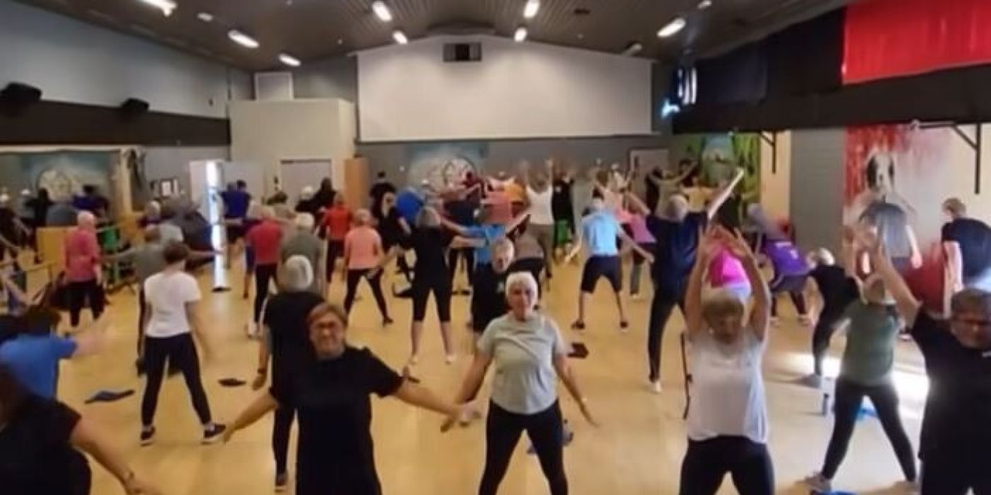 En stor gruppetrening i en romslig innendørs treningssal. Rommet er fylt med mennesker, hovedsakelig eldre voksne, som deltar i en aerobic-økt.