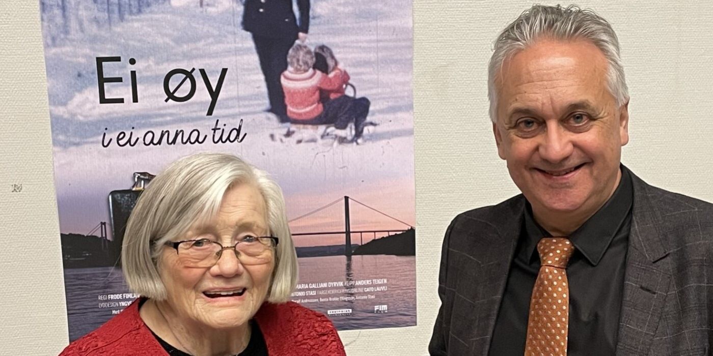 Bildet viser to personer stående foran en plakat med tittelen "Ei øy i ei anna tid". Personen til venstre er en eldre kvinne med grått hår og briller, iført en rød topp. Personen til høyre er en mann med grått hår, iført dressjakke, sort skjorte og oransje slips med mønster. Begge smiler mot kameraet. Bakgrunnen domineres av filmplakaten, som viser en vinterlig scene med en person som trekker et barn på akebrett.