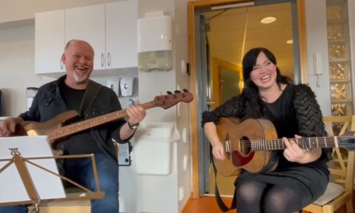 Bildet viser Berit Håpoldøy som ler og holder en gitar i hånden. Ved siden av seg sitter Bjørn Tore Bystrøm. Han spiller bassgitar og er også i godt humør.